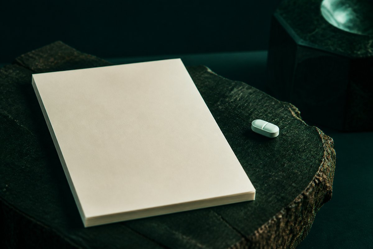 Forest-green still life: unsigned cream prescription pad and one pale tablet on a forest slab